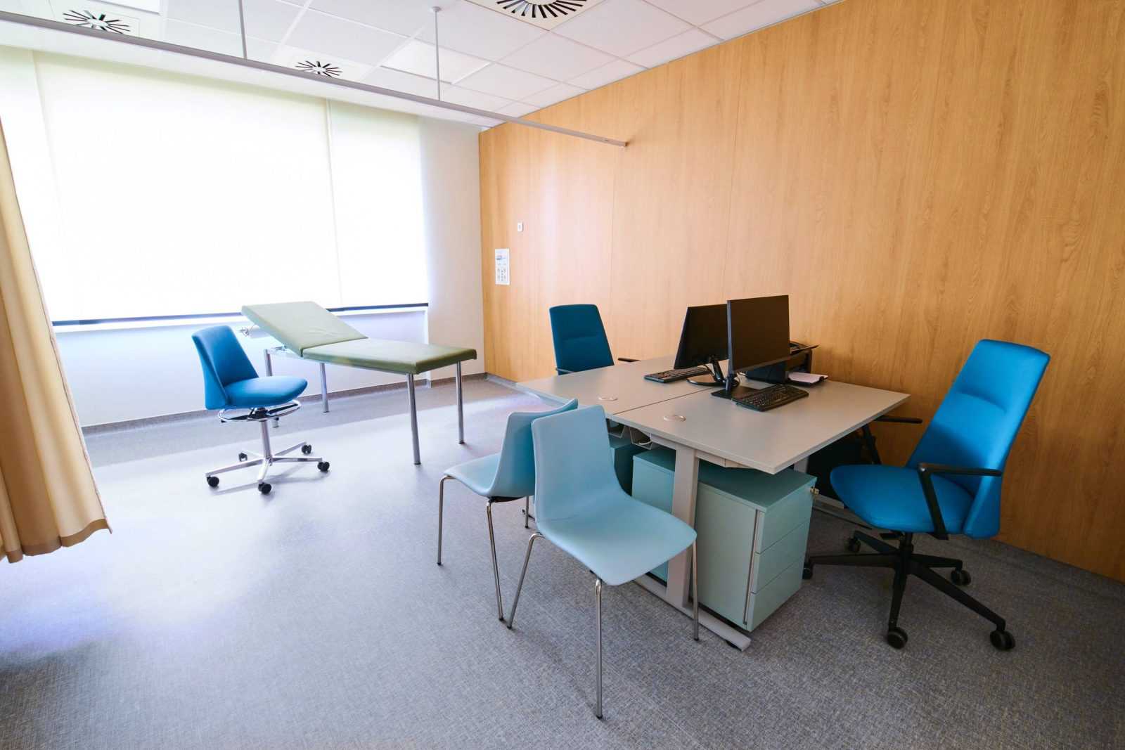 Modern medical examination room with blue chairs and exam table.