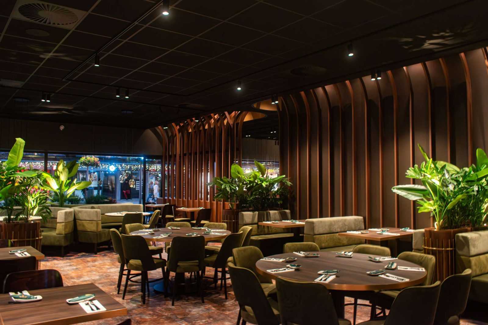Modern restaurant interior with wooden tables, green chairs, and tropical plants.