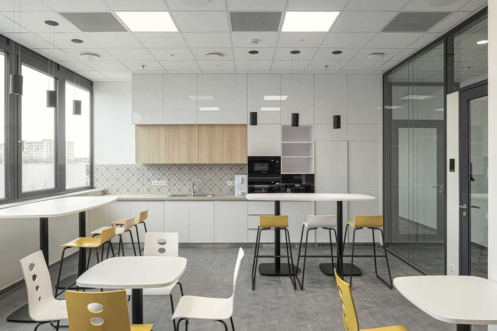 Modern office kitchen with white cabinets, yellow chairs, and large windows.