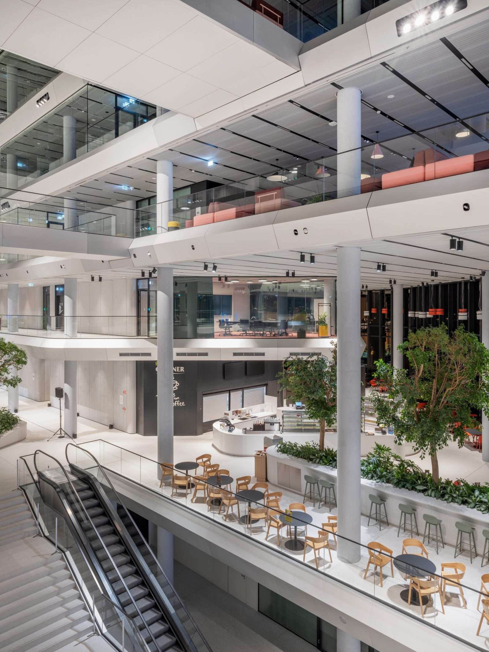Modern multi-level office atrium with glass railings, escalator, and indoor greenery.