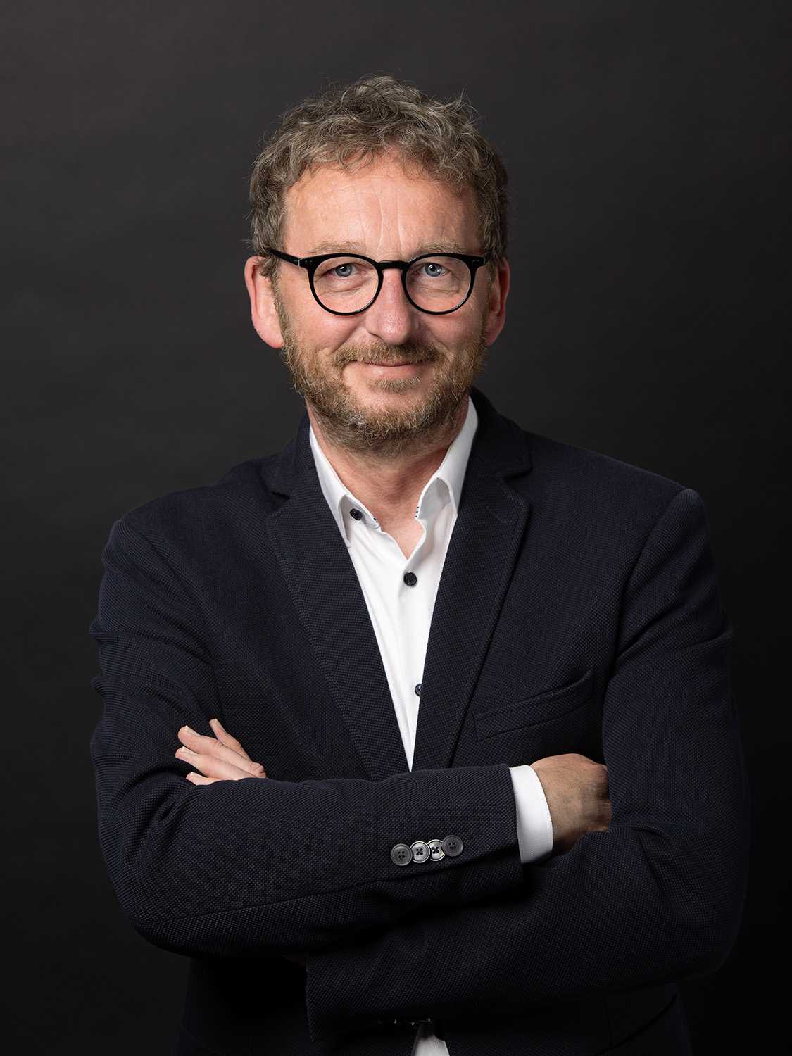 Professional headshot of a man with glasses and beard wearing a dark blazer.